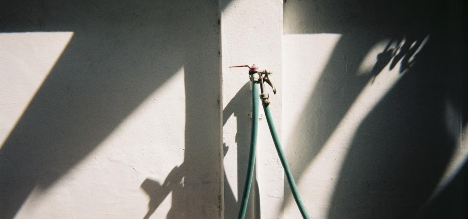 Two green garden hoses still attached to an outdoor faucet on a sunlit white wall